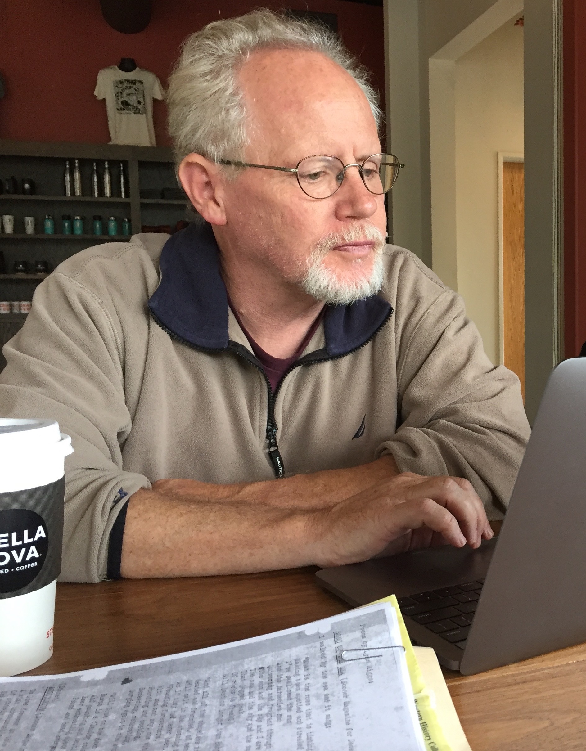 Crag Hill has short gray hair and glasses and is focused on a laptop in a cozy café setting. He is wearing a beige collared pullover. A coffee cup and papers are on the wooden table were he sits, suggesting a relaxed work atmosphere.